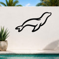Black metal sea lion wall art with a sleek silhouette design, mounted on an outdoor white wall above a pool with palm trees in the background.