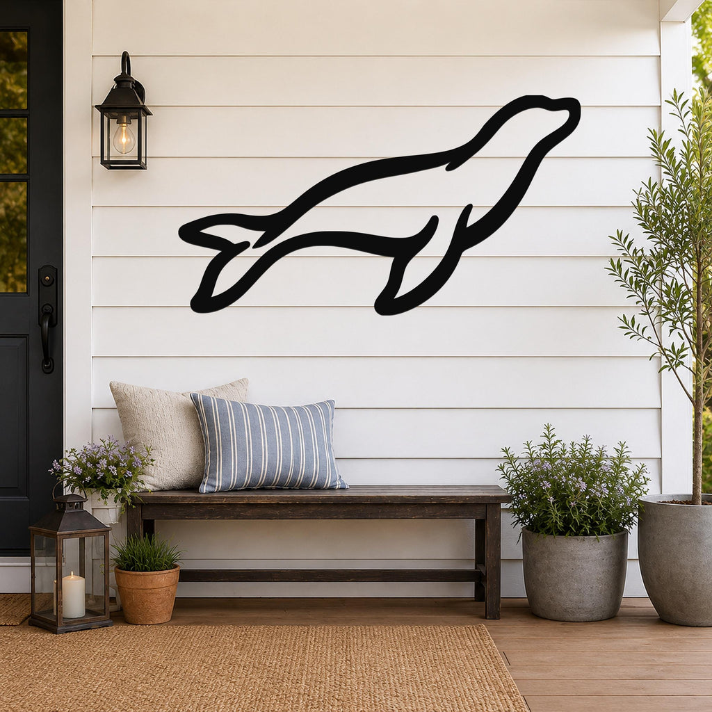 Black metal sea lion wall art silhouette mounted on a white shiplap wall above a wooden bench on a porch.
