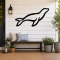 Black metal sea lion wall art silhouette mounted on a white shiplap wall above a wooden bench on a porch.