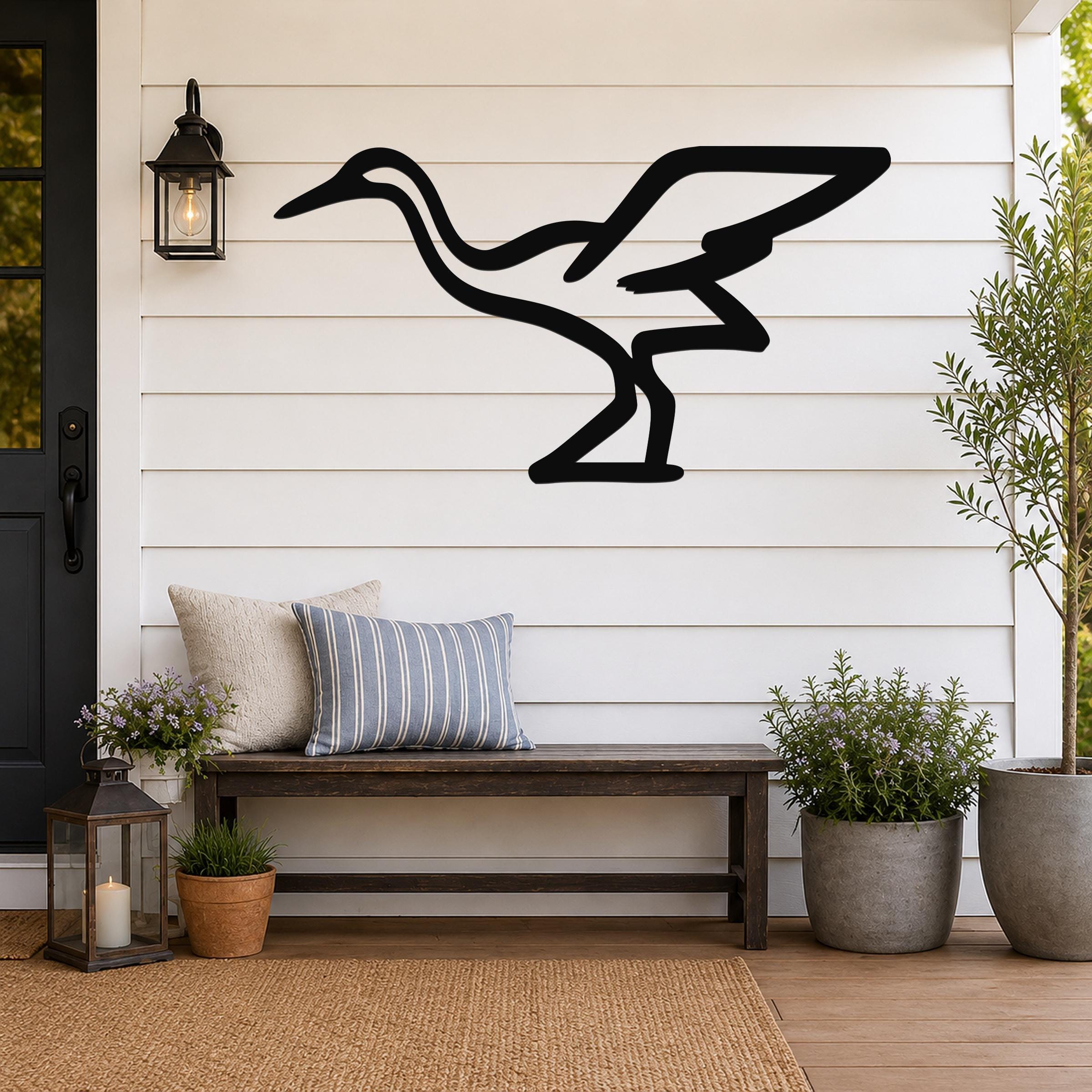 Black metal crane wall art silhouette mounted on a white shiplap wall above a wooden bench on a porch.