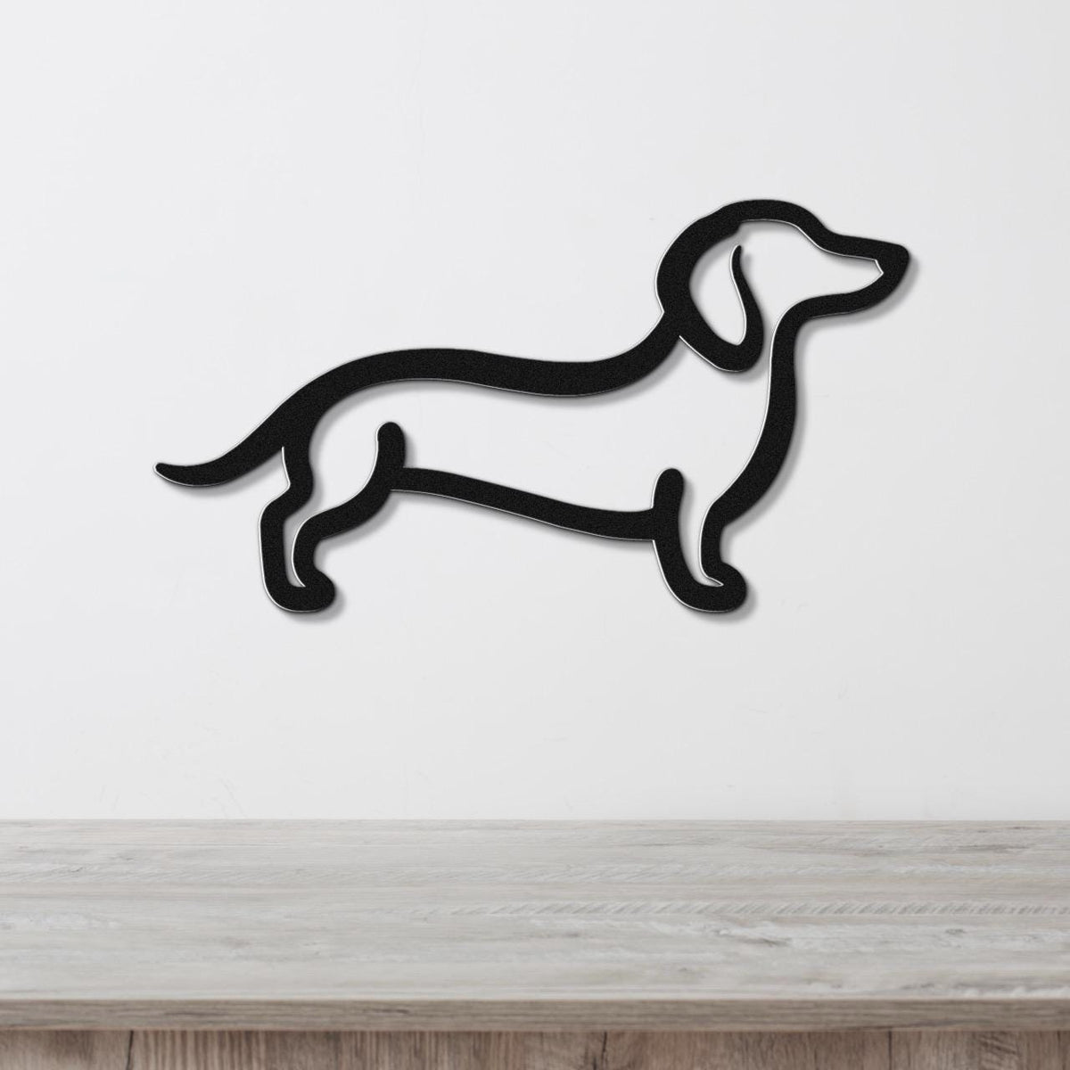 Black metal dachshund wall art in a silhouette cutout style, mounted on a white wall above a light wood surface.