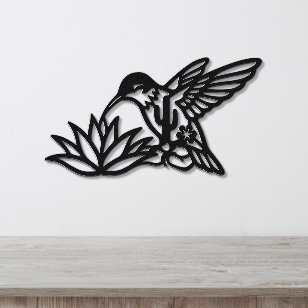 Black metal hummingbird wall art with cutout wings and floral details, mounted on a white wall above a light wood surface.