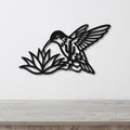 Black metal hummingbird wall art with cutout wings and floral details, mounted on a white wall above a light wood surface.