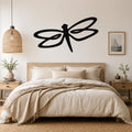 Black metal dragonfly wall art with flowing wing cutouts, mounted above a wooden bed in a neutral bedroom.