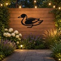 Black metal duck wall sculpture with a silhouette cutout, mounted on a wooden fence in an outdoor garden setting with string lights and plants.