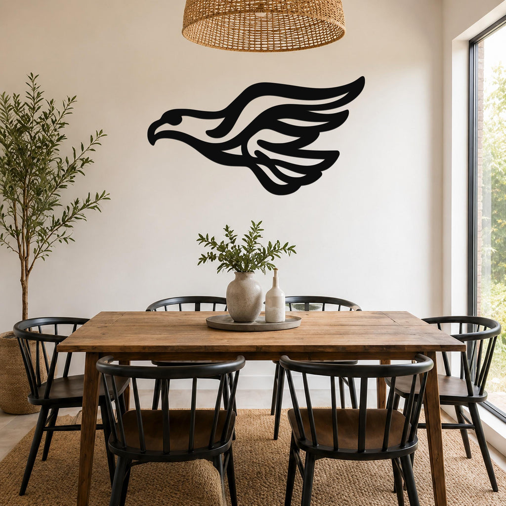 Black metal eagle wall art with flowing wing cutouts, mounted on a white wall above a wooden dining table in a bright dining room.