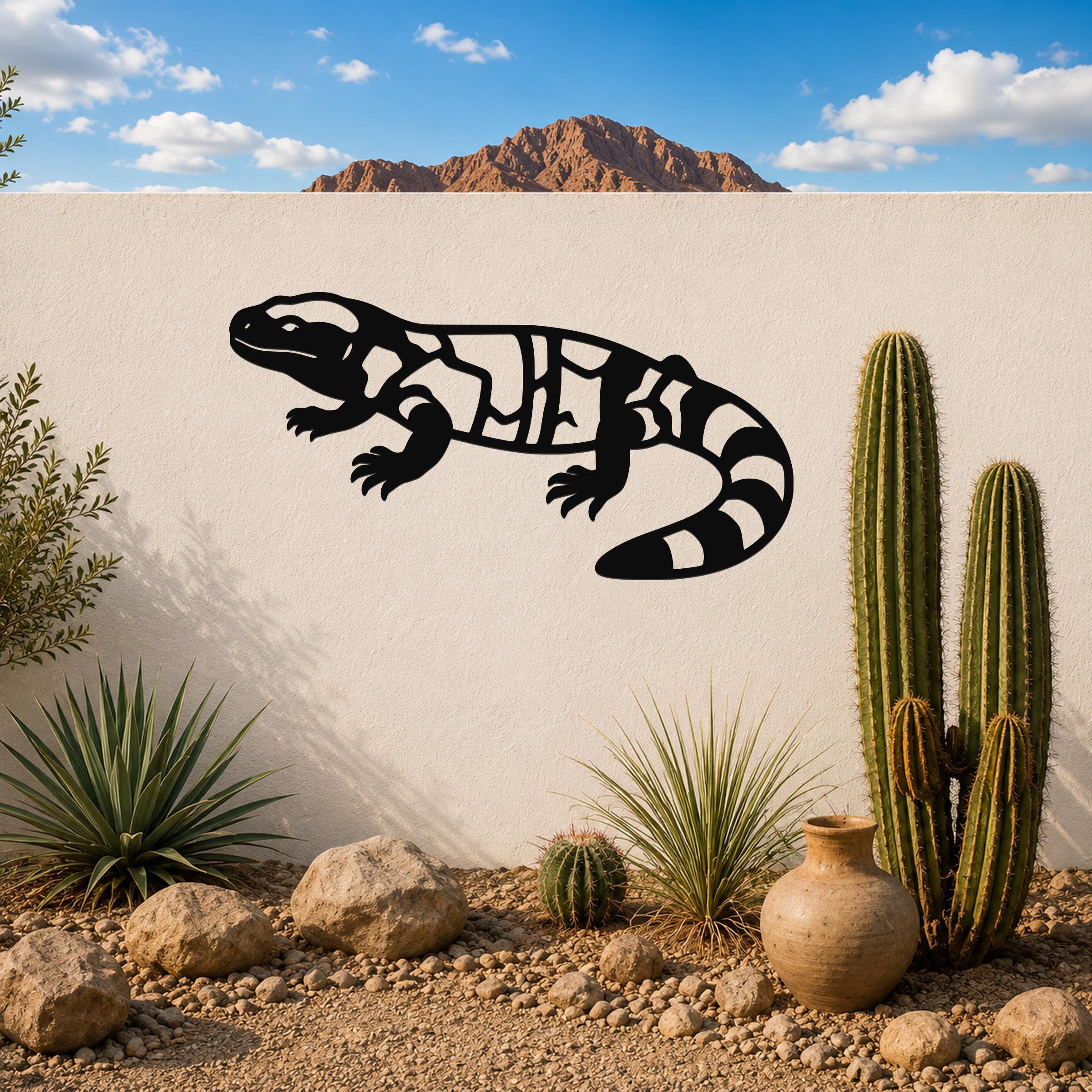 Black metal Gila monster wall art with internal cutout patterns, mounted on a white outdoor wall above a desert garden with cacti and rocks.