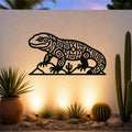 Black metal wall art of a Gila monster with intricate geometric cutouts, mounted on an outdoor wall above a desert garden with cacti and warm uplighting.
