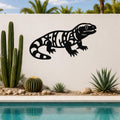 Black metal wall art of a Gila monster with intricate internal cutouts, mounted on a white outdoor wall above a pool with cacti and desert plants.