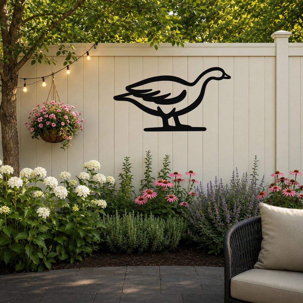 Black metal goose wall art silhouette mounted on a light beige outdoor fence, above a garden bed with white and pink flowers and a hanging basket, on a patio with string lights.