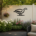 Black metal goose wall art silhouette mounted on a light beige outdoor fence, above a garden bed with white and pink flowers and a hanging basket, on a patio with string lights.