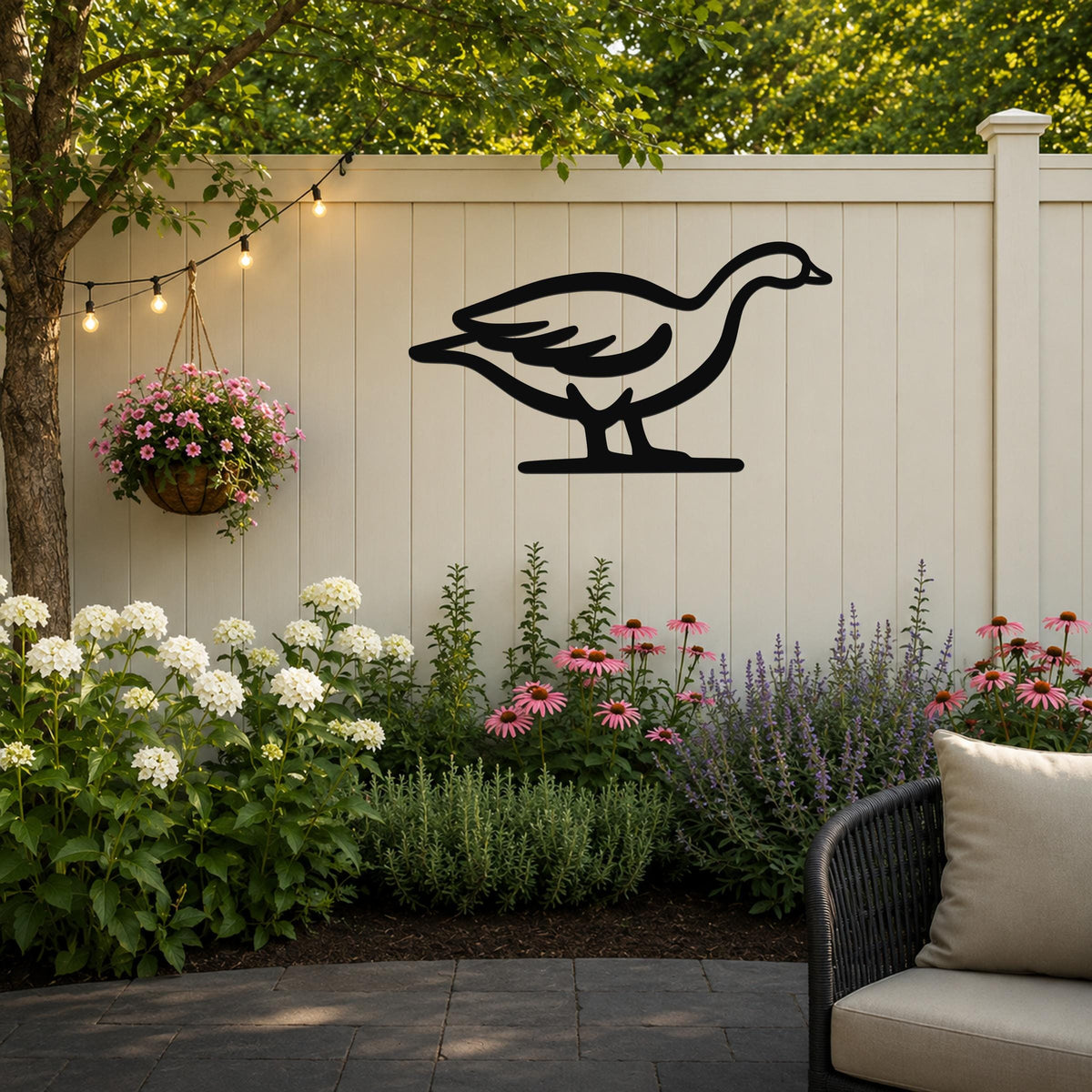 Black metal goose wall art silhouette mounted on a light beige outdoor fence, above a garden bed with white and pink flowers and a hanging basket, on a patio with string lights.