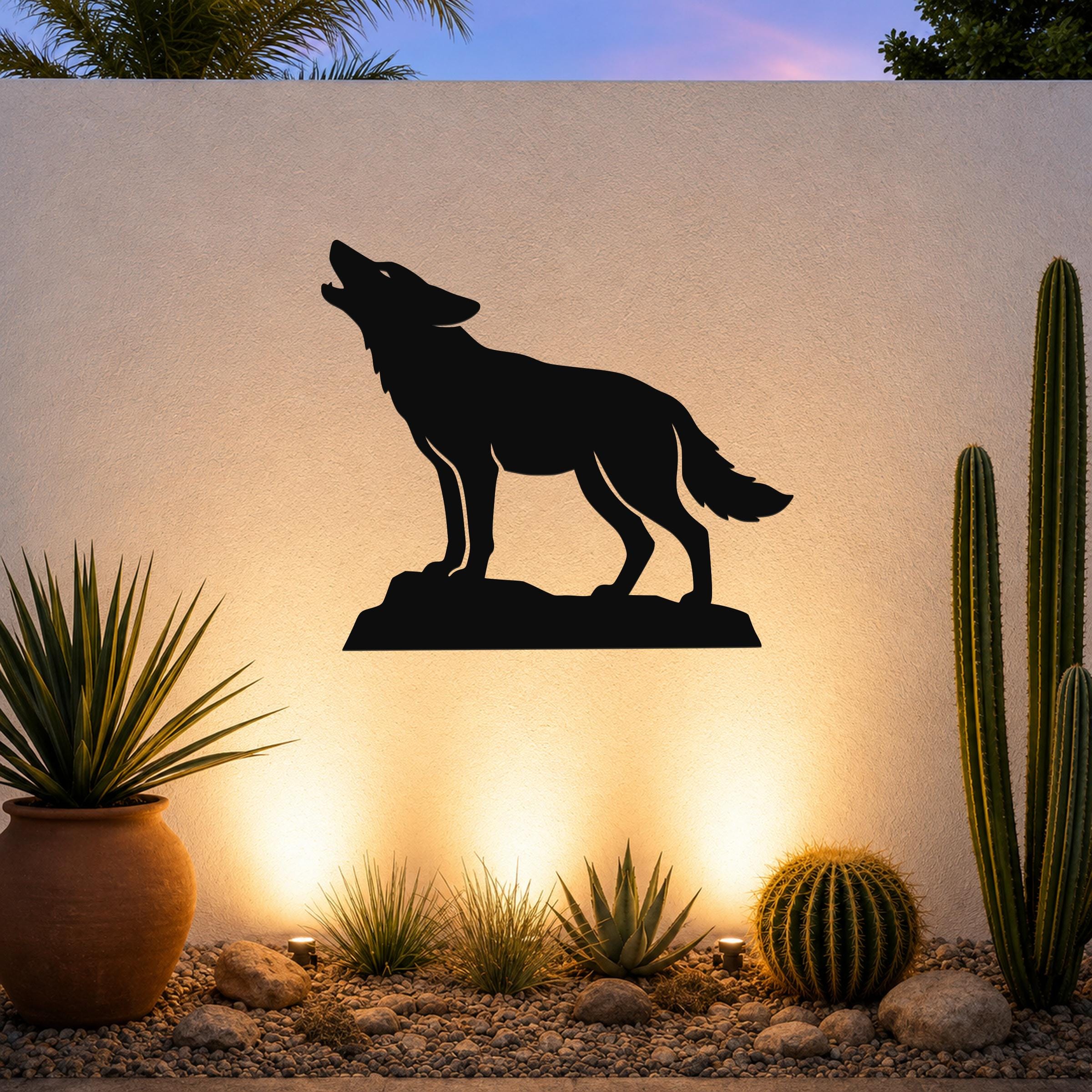 Black metal coyote wall art silhouette howling on a rock, mounted on an outdoor wall above a desert garden with cacti and warm uplighting.