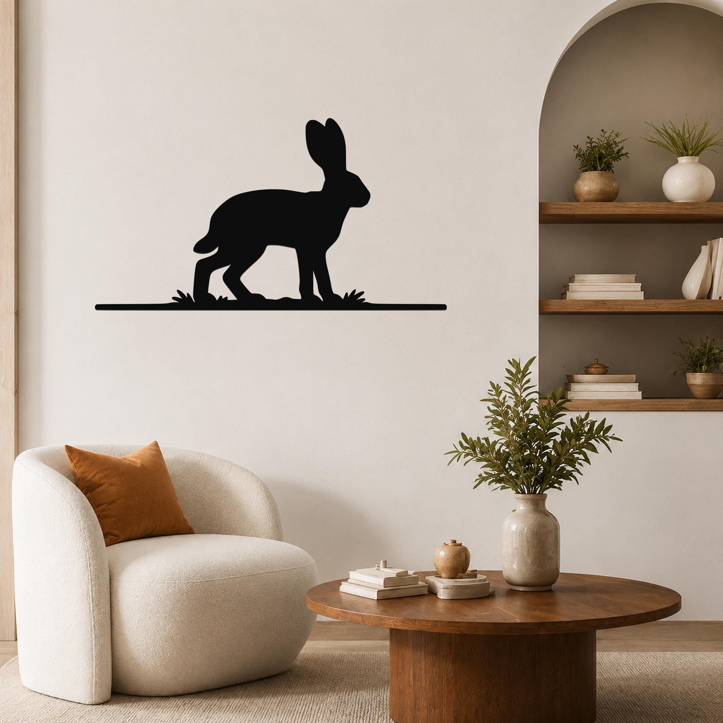 Black metal jackrabbit wall art silhouette mounted on a living room wall above a white armchair, with a wooden coffee table and built-in shelves visible nearby.
