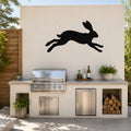 Black metal jackrabbit wall art silhouette mounted on a light-colored outdoor wall above a kitchen counter with a grill and plants.