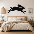 Black metal jackrabbit wall art silhouette with motion lines, mounted above a bed in a neutral bedroom.