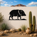 Black metal javelina wall art silhouette with white facial and neck details, mounted on a white outdoor wall above a desert garden with cacti and rocks.