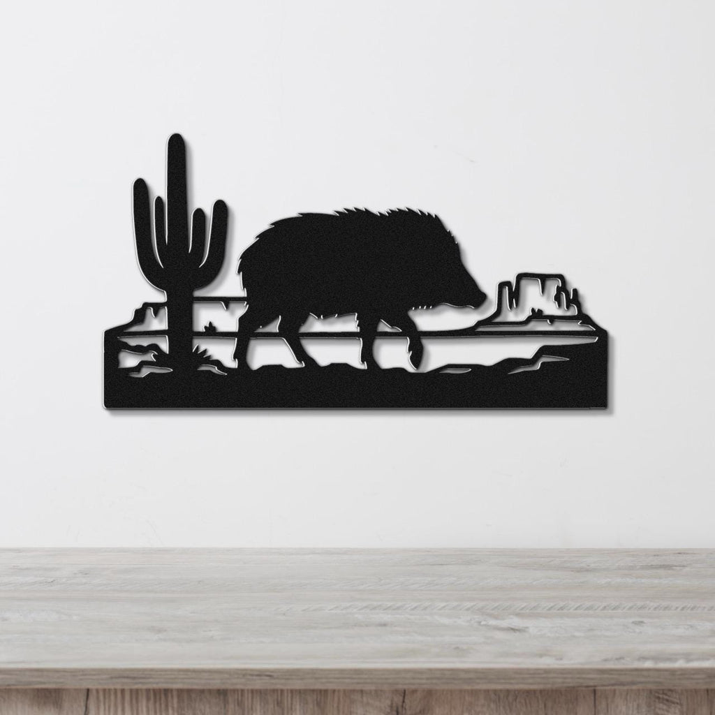 Black metal wall art featuring a javelina silhouette walking past a saguaro cactus and canyon rock formations, mounted on a white wall above a wooden surface.