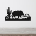 Black metal wall art featuring a javelina silhouette walking past a saguaro cactus and canyon rock formations, mounted on a white wall above a wooden surface.