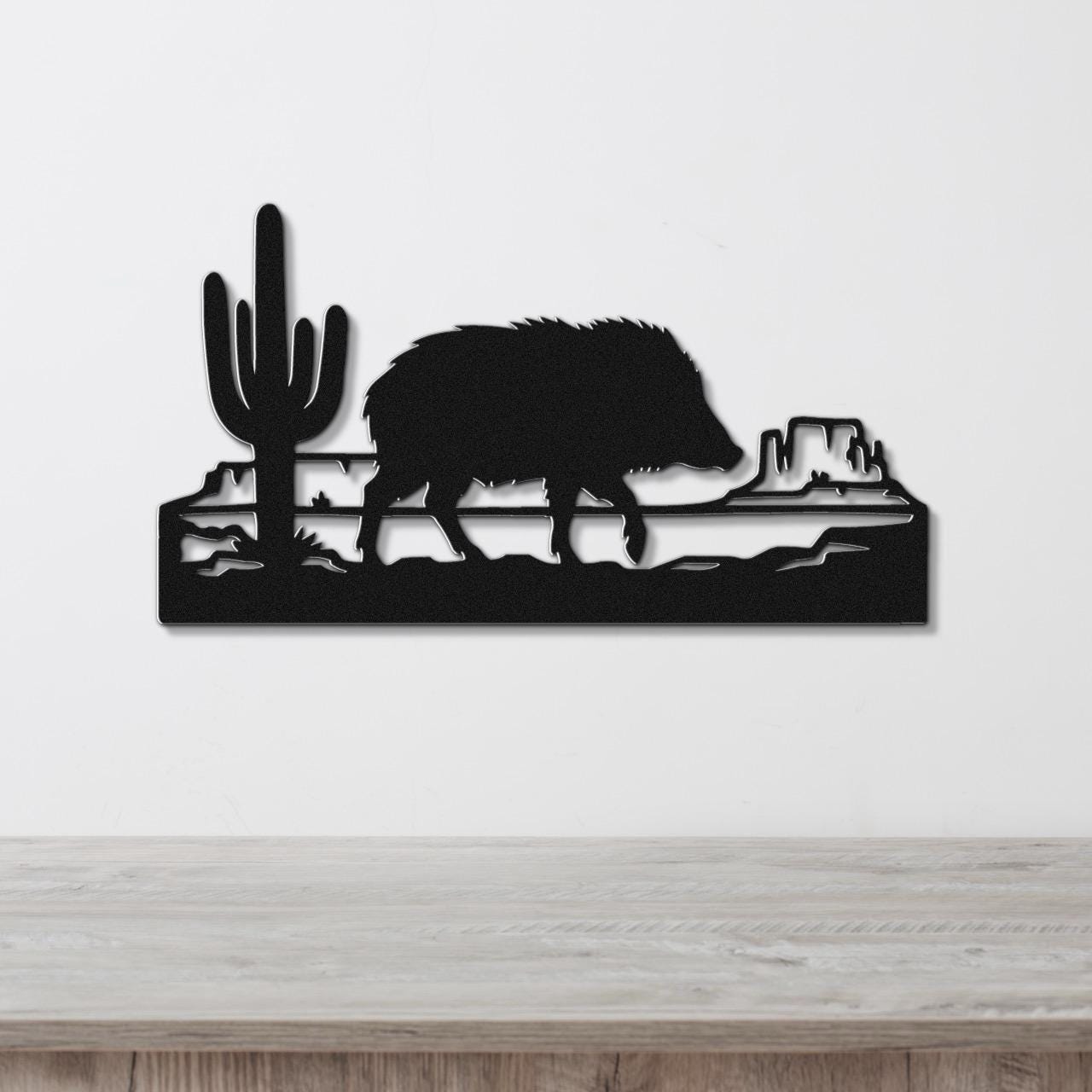 Black metal wall art featuring a javelina silhouette walking past a saguaro cactus and canyon rock formations, mounted on a white wall above a wooden surface.