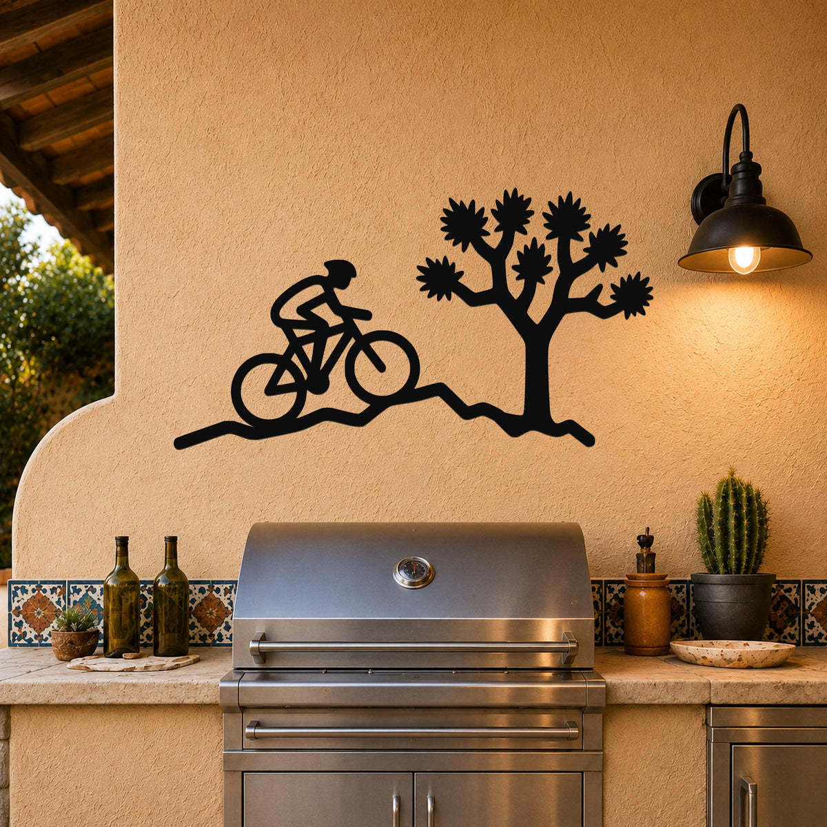 Black metal wall art cutout of a cyclist on a trail beside a Joshua tree, mounted on a stucco wall above a stainless steel grill in an outdoor kitchen.