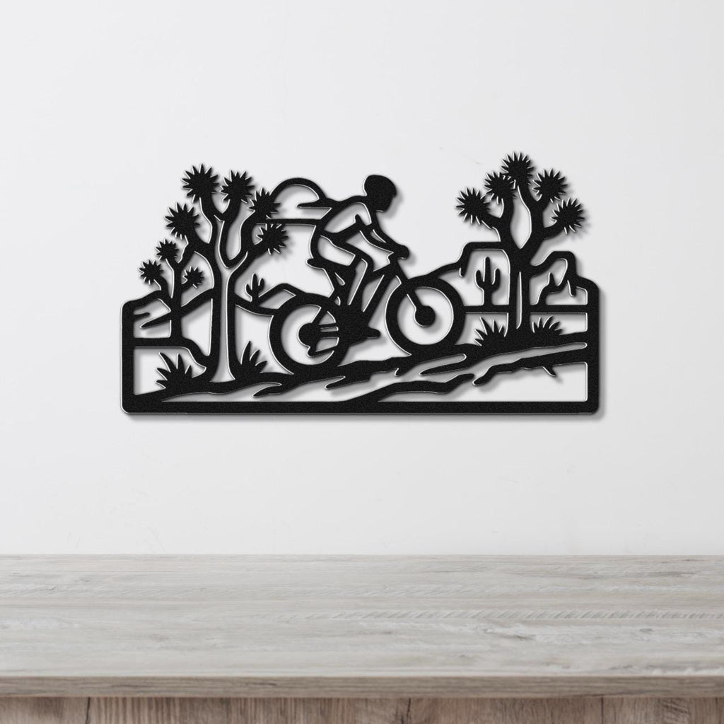 Black metal wall art cutout of a cyclist riding through a desert landscape with Joshua trees and cacti, mounted on a white wall above a wooden surface.