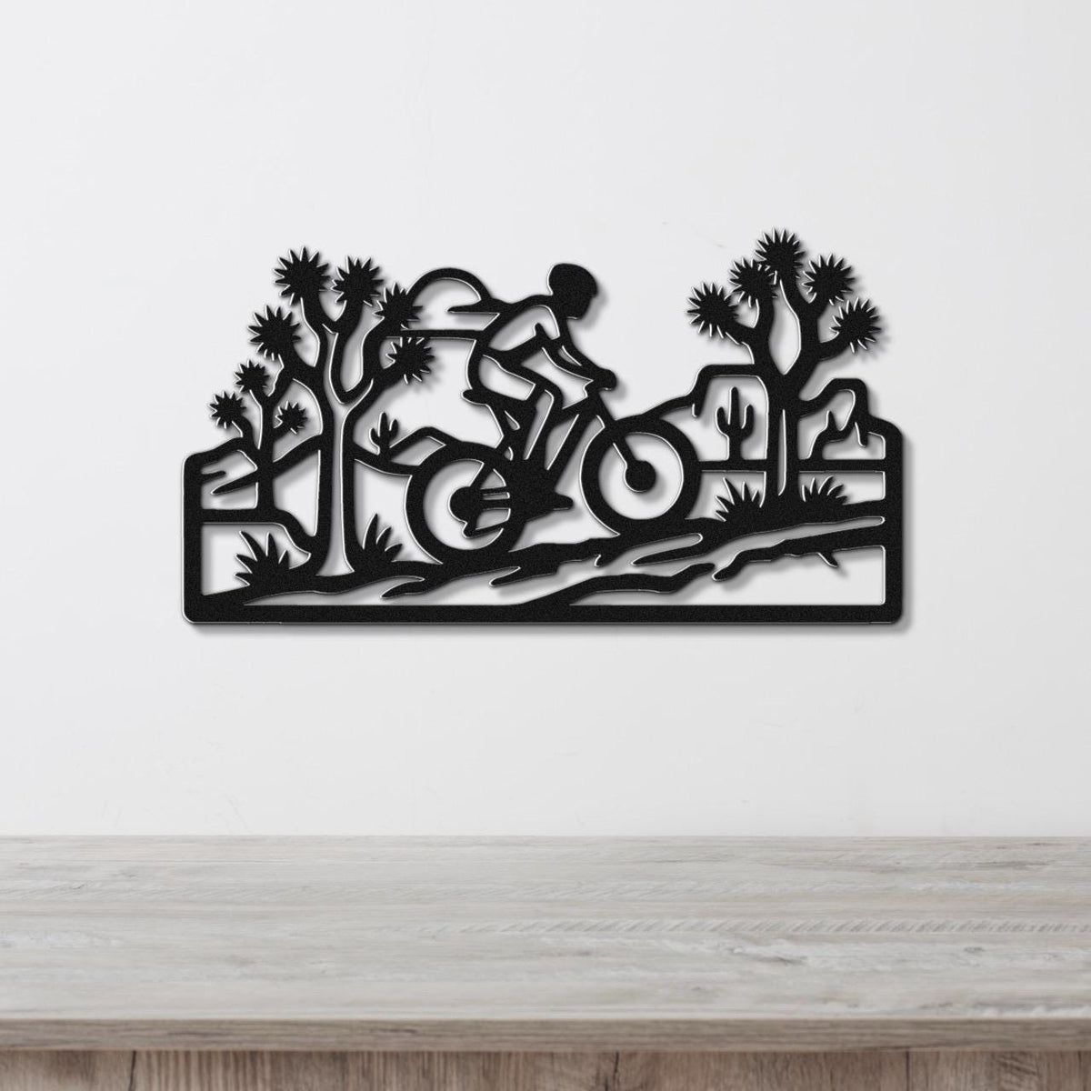 Black metal wall art cutout of a cyclist riding through a desert landscape with Joshua trees and cacti, mounted on a white wall above a wooden surface.