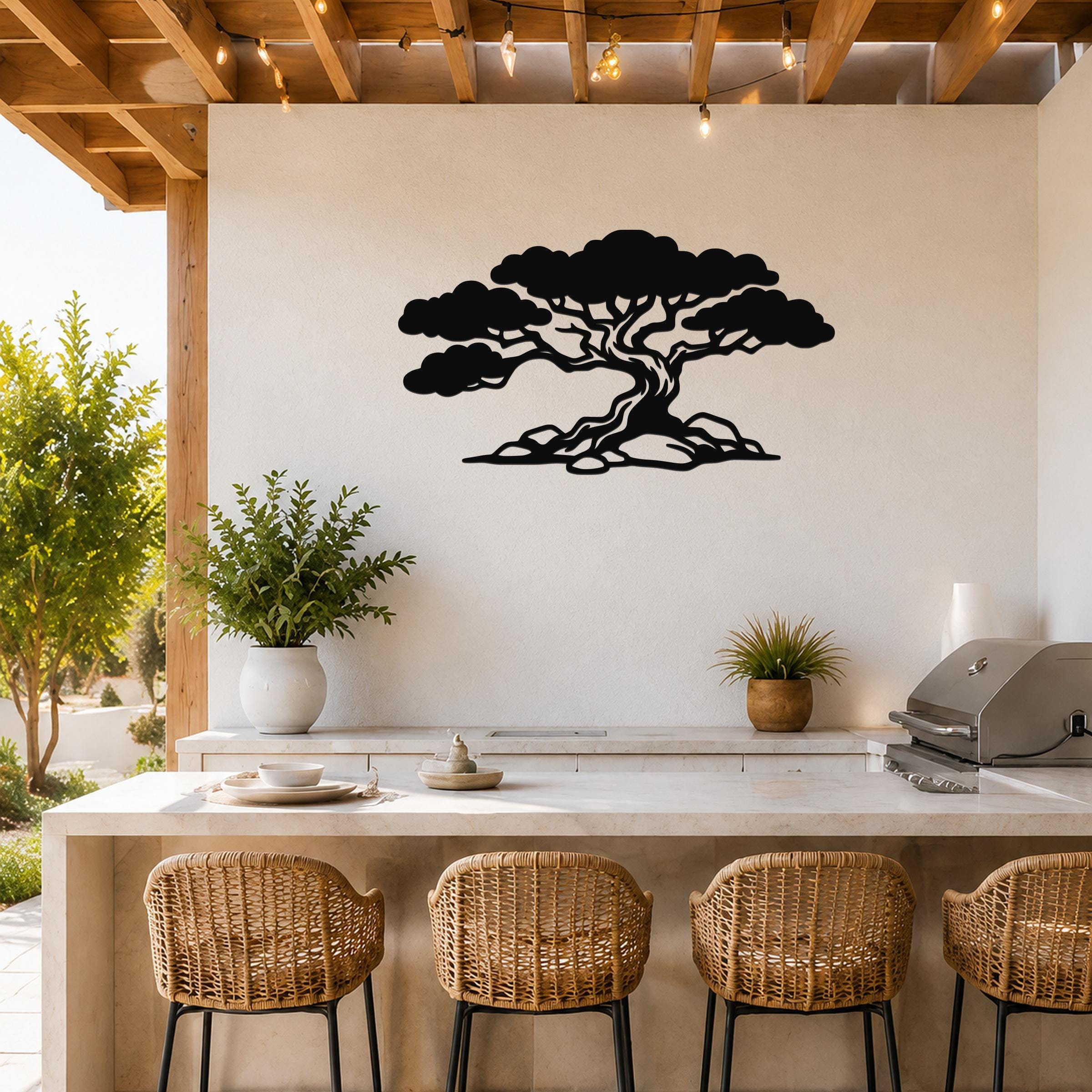 Black metal wall art of a wind-sculpted juniper tree silhouette, mounted on a white outdoor wall above a kitchen counter with wicker bar stools.