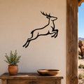 Black metal jumping deer wall art with motion-line cutouts, mounted on a beige wall above a wooden table in a rustic outdoor entryway.