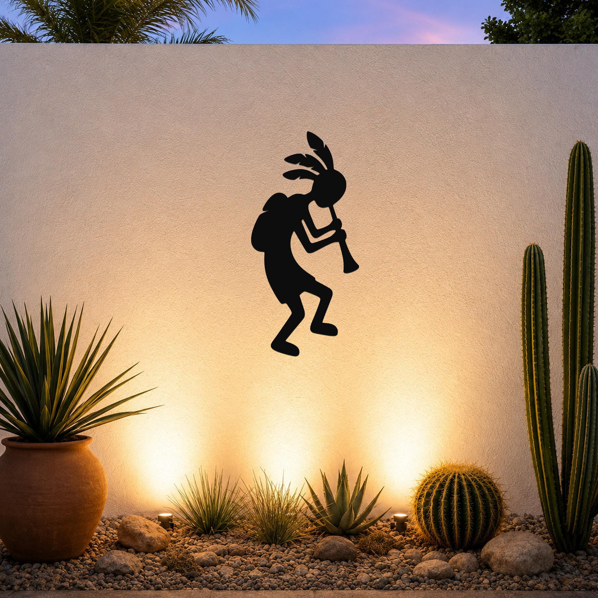 Black metal Kokopelli wall art silhouette playing a flute, mounted on a white outdoor wall above a desert garden with cacti and warm uplighting.