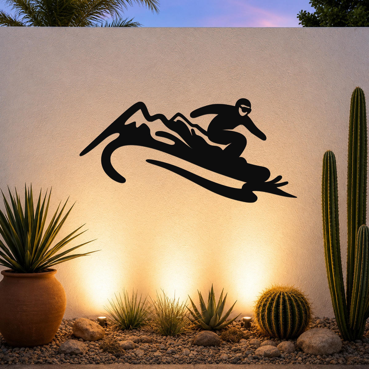 Black metal wall art silhouette of a snowboarder in motion, mounted on an outdoor wall above a desert garden with cacti and warm ground lighting.