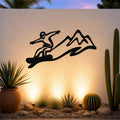 Black metal snowboarder wall art with mountain silhouette, mounted on an outdoor wall above a desert garden with cacti and warm uplighting.