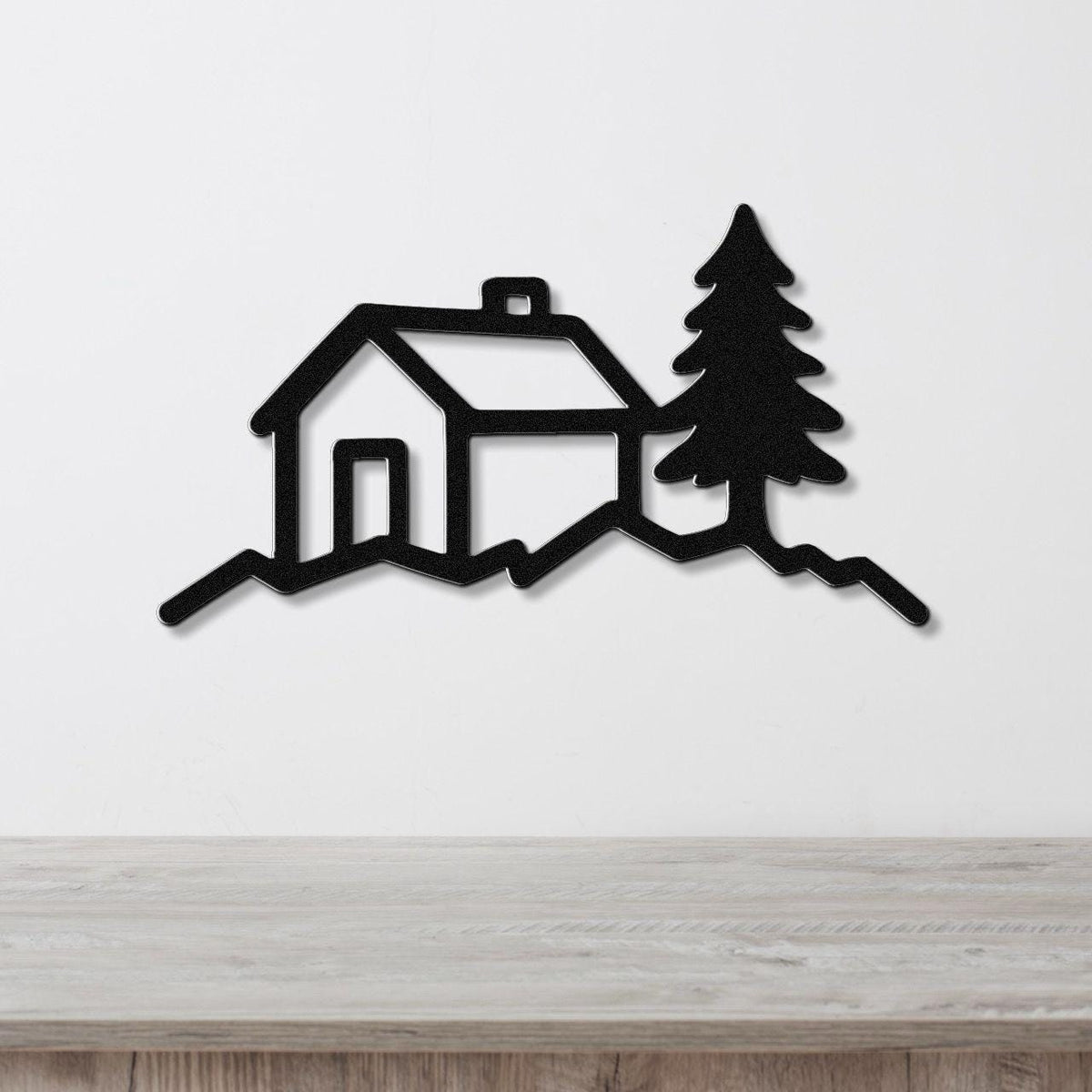 Black metal wall art cutout of a cabin and pine tree silhouette, mounted on a white wall above a light wood counter.
