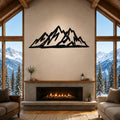 Black metal mountain silhouette wall art mounted above a fireplace in a rustic living room with large windows overlooking snowy peaks.