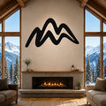 Black metal mountain silhouette wall art mounted above a fireplace in a rustic living room with large windows overlooking snowy peaks.