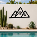 Black metal mountain silhouette wall art with layered peaks, mounted on a white outdoor wall above a pool with cacti and palm trees in the background.