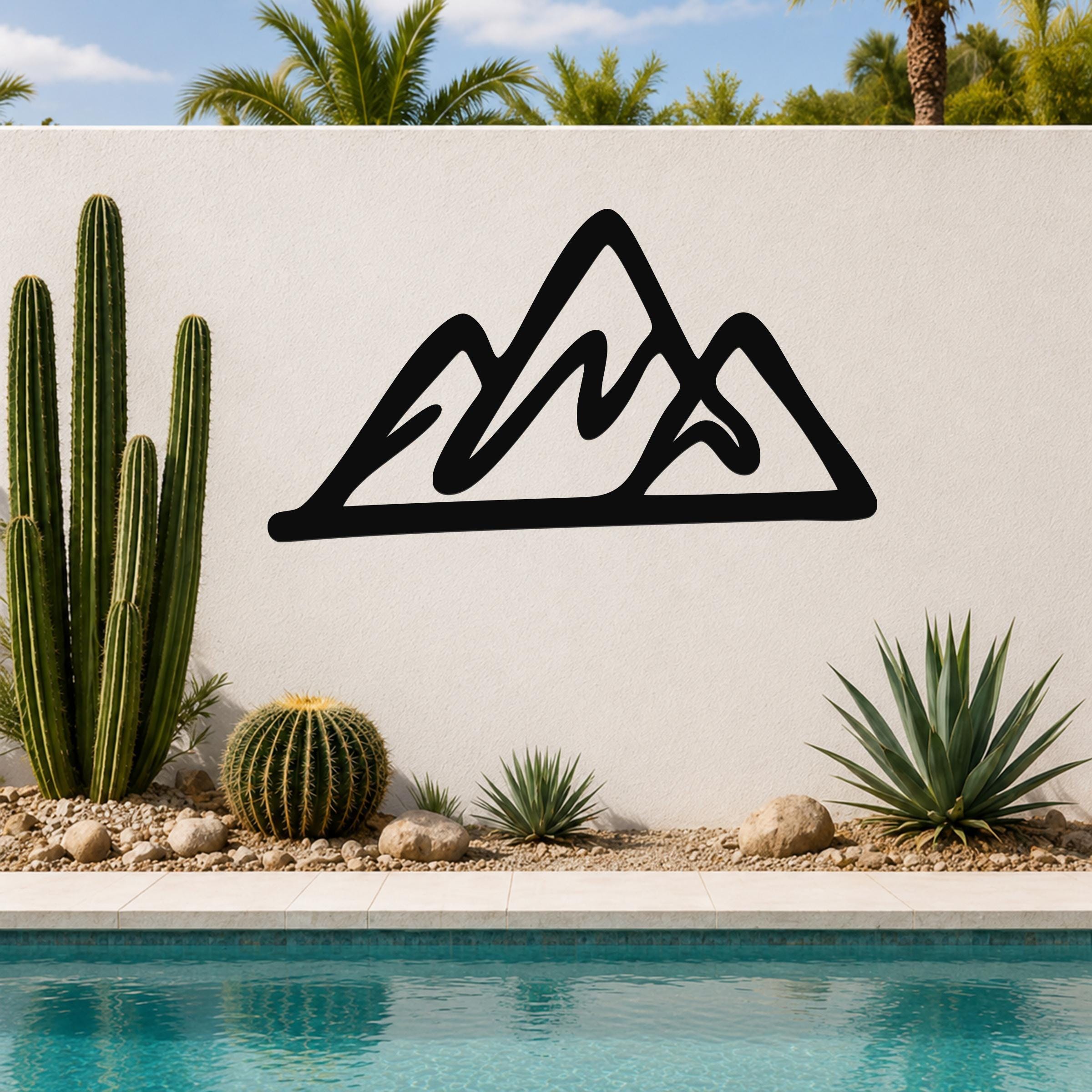 Black metal mountain silhouette wall art with layered peaks, mounted on a white outdoor wall above a pool with cacti and palm trees in the background.