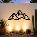 Black metal mountain range wall art with a silhouette design, mounted on an outdoor wall above a desert garden with cacti and warm ground lighting.