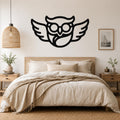 Black metal wall art silhouette of a bespectacled owl with spread wings, mounted above a bed in a neutral bedroom.