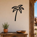 Black metal palm tree wall art with a silhouette design, mounted on a beige wall above a wooden table in a patio setting.