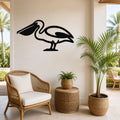 Black metal pelican wall art silhouette mounted on a beige wall above a wicker chair in a coastal patio setting.