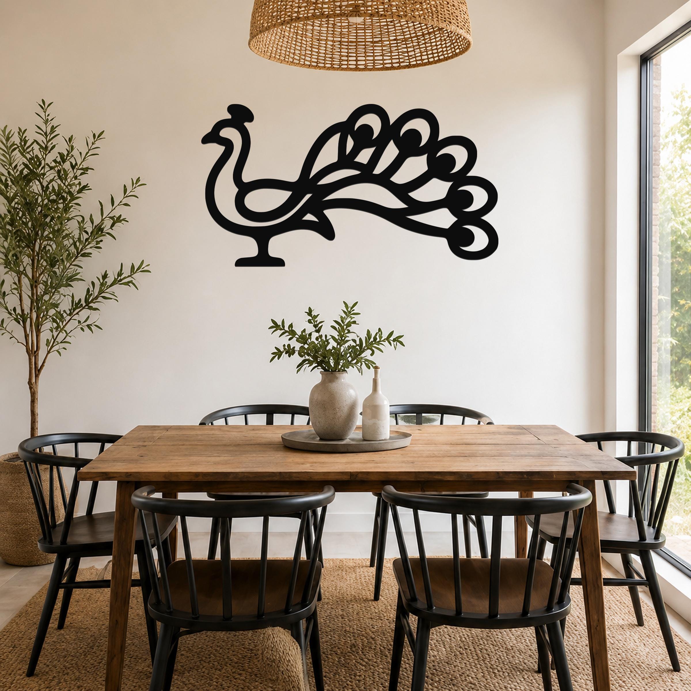 Black metal peacock wall art with a cutout silhouette of a fanned tail, mounted on a white wall above a wooden dining table in a bright dining room.