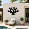 Black metal prickly pear cactus wall art with flower and spine cutouts, mounted on a white outdoor wall above a lounge chair near a pool.