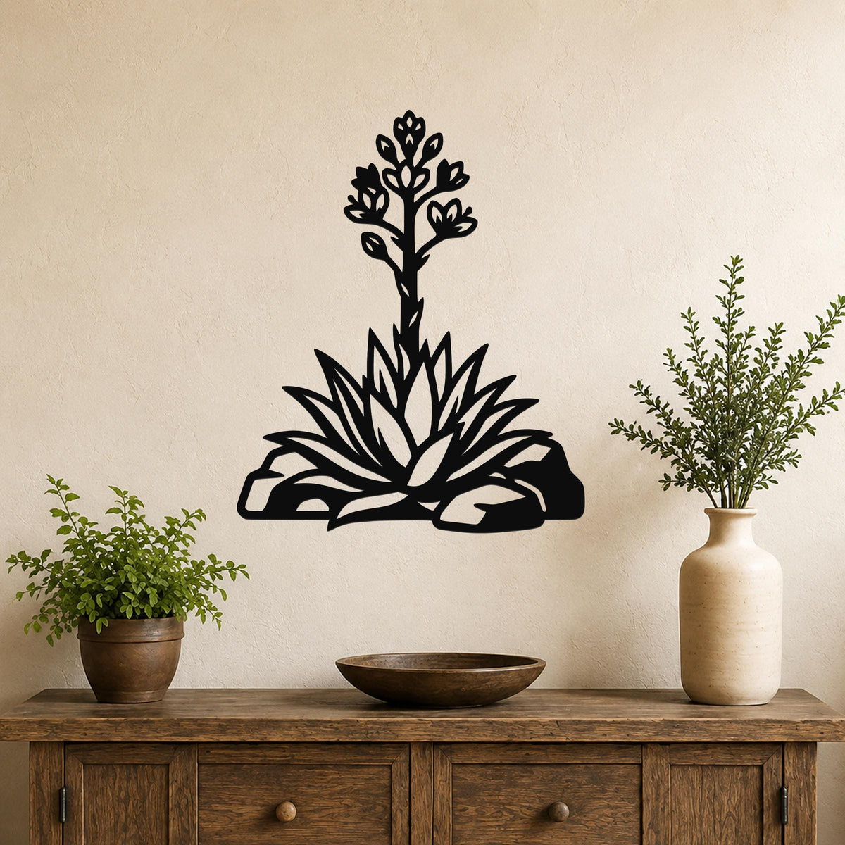 Black metal agave wall art with blooming flower cutout, mounted on a beige wall above a wooden console table with potted plants.