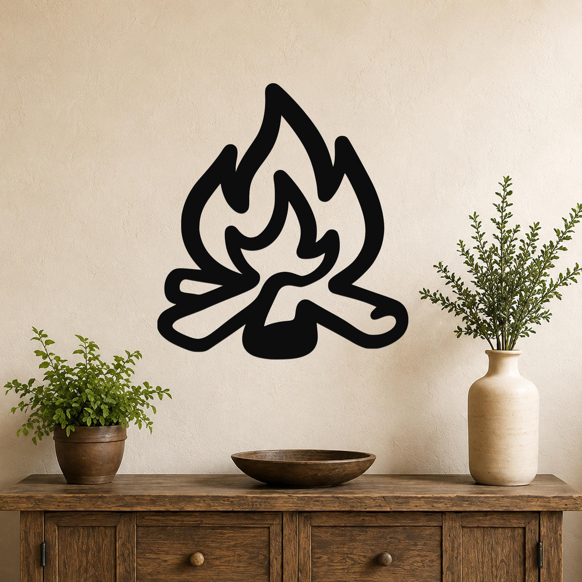 Black metal campfire wall art silhouette with flame and log cutouts, mounted on a beige wall above a wooden console table with potted plants.