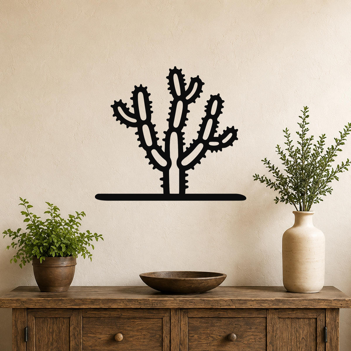 Black metal cholla cactus wall art with spiny branch cutouts, mounted on a beige wall above a wooden console table with potted plants.