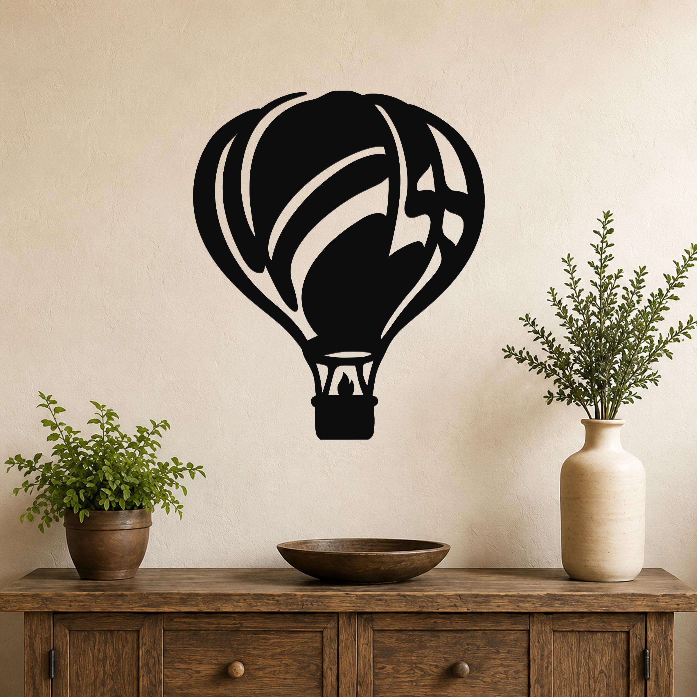 Black metal hot air balloon wall art with internal flame and basket cutouts, mounted on a beige wall above a wooden console table with plants.