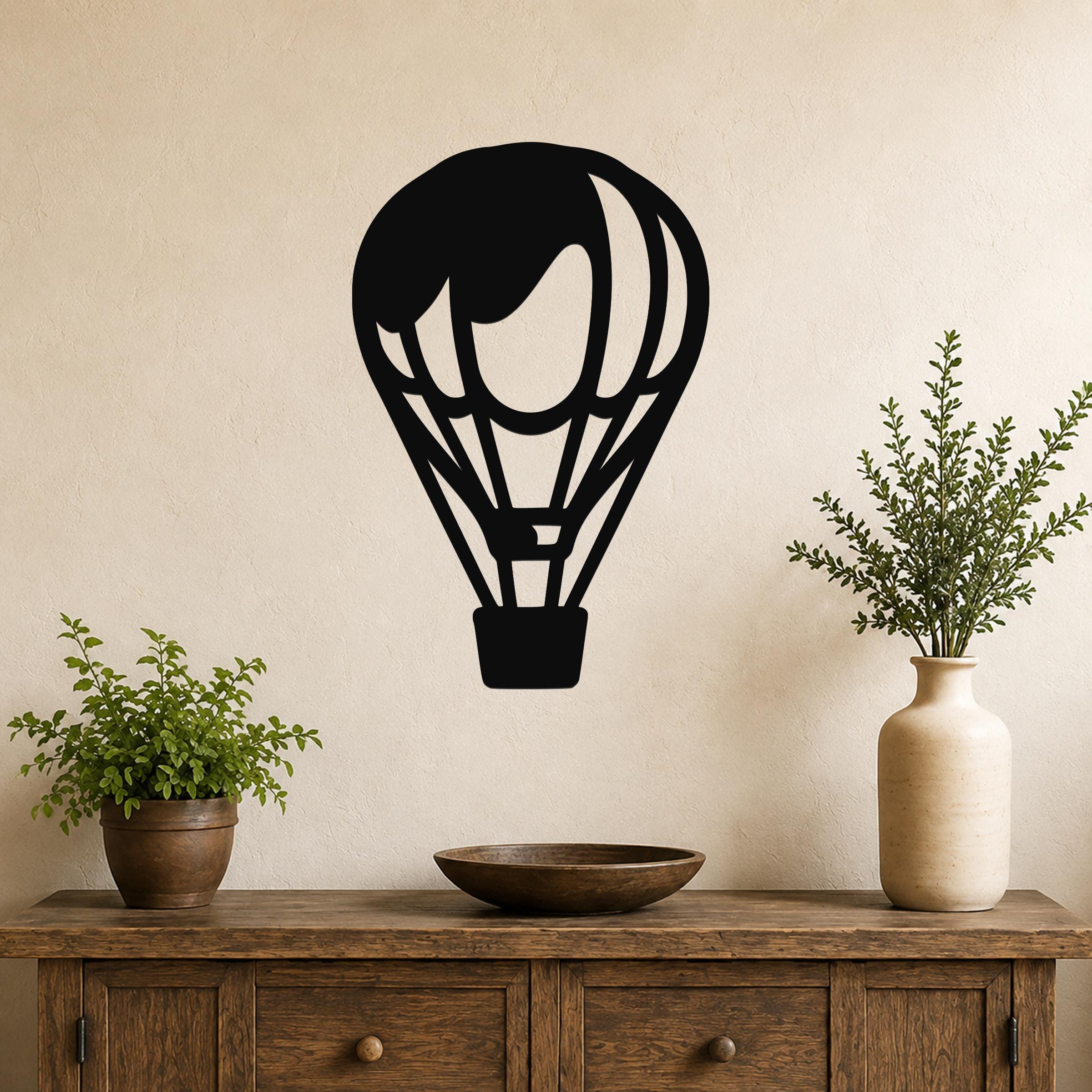 Black metal hot air balloon wall art with internal cutout details, mounted on a beige wall above a wooden console table with potted plants.