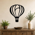 Black metal hot air balloon wall art with internal flame and basket cutouts, mounted on a beige wall above a wooden console table with plants.