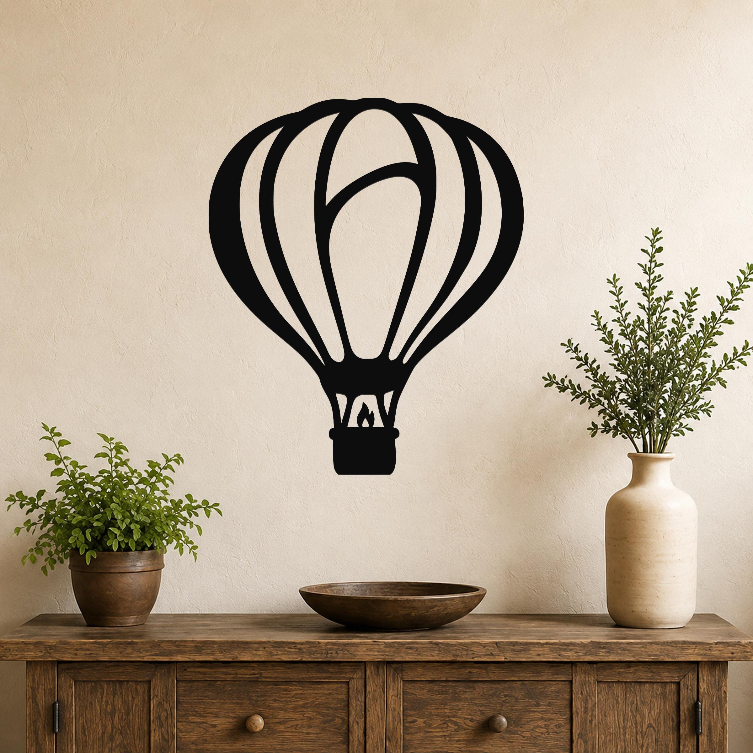 Black metal hot air balloon wall art with internal flame and basket cutouts, mounted on a beige wall above a wooden console table with plants.